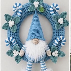 Op de afbeelding: Een blauwe gehaakte krans met een centrale kabouter met een hoge blauwe muts, een witte baard van imitatiebont en gestreepte benen. De krans is versierd met gehaakte groene bladeren, witte bessen en blauw-wit gestreepte zuurstokken.