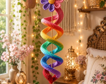 Op de afbeelding: Een hangende haakdecoratie met een grote roze en paarse bloem aan de bovenkant, gevolgd door een lange, spiraalvormige, regenboogkleurige buis van garen. Een kleine, lichtblauwe, druppelvormige haakbedel hangt aan de onderkant van de spiraal.