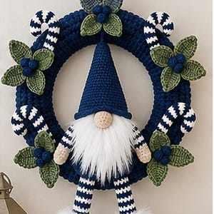 Op de afbeelding: Een donkerblauwe gehaakte krans met een centrale kabouter met een hoge blauwe muts, witte baard en gestreepte benen. De krans is versierd met groene gehaakte bladeren, donkerblauwe bessen en blauw-wit gestreepte zuurstokken.