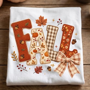 Puede incluir: Una camiseta blanca con la palabra FALL bordada en letras grandes y decorativas. Las letras están rellenas con patrones de calabazas, hojas y bellotas en tonos naranja, marrón y crema. Un lazo de tela a cuadros está sujeto a la letra L.