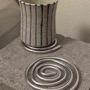 May include: A ceramic cup with a speckled off-white glaze and vertical dark blue stripes, resting on a silver-toned metal spiral coaster. A second identical silver metal spiral coaster is positioned in front of the cup on the surface.