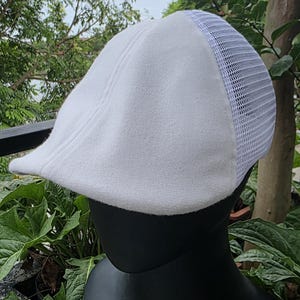 May include: A white flat cap featuring a solid fabric front panel and a breathable white mesh back panel. The cap is displayed on a black mannequin head, showcasing its structured shape, curved brim, and the contrast between the smooth fabric and mesh.