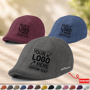 Puede incluir: Tres gorras planas en color burdeos, azul y gris, cada una con un diseño impreso en negro que dice YOUR LOGO HERE CUSTOM TEXT. La gorra gris también incluye el nombre Williams bordado en blanco en el lateral, con una flecha roja señalándolo.