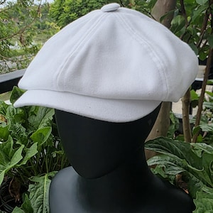 Puede incluir: Una gorra de estilo newsboy en color blanco sólido que presenta una corona redondeada de varios paneles con un pequeño botón cubierto de tela en la parte superior central y una visera corta y rígida al frente. Exhibida sobre un maniquí negro.