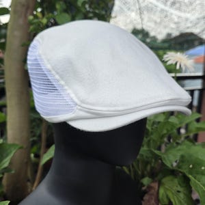 Puede incluir: Una gorra plana de color blanco que presenta una corona de tela sólida y un panel de malla transpirable en el lateral. La gorra tiene una visera corta y rígida, con una forma estructurada diseñada para ajustarse a la cabeza en color blanco.