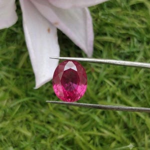 Peut inclure: Une pierre précieuse ovale aux reflets magenta-rose profond et à la surface à facettes, maintenue par une pince métallique argentée. La pierre présente une grande clarté et une réflexion lumineuse sur ses facettes polies et symétriques.
