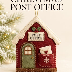 Könnte beinhalten: Ein Filzornament in Hausform mit dem Text "WOODLAND CHRISTMAS POST OFFICE" und "POST OFFICE" auf dem Gebäude. Es verfügt über eine rote Fassade, eine grüne Tür, ein weißes Dach und eine Tasche mit einem kleinen Umschlag mit einem roten Herz.