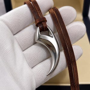 Puede incluir: Un colgante de metal plateado con forma de garra o diente curvado, con un acabado liso y pulido. El colgante está suspendido de un cordón de cuero marrón de doble hebra que pasa a través de un bucle de metal en la parte superior del diseño.