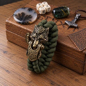 May include: An olive green paracord bracelet featuring a metallic bronze-toned wolf head clasp with intricate engraved details. The bracelet is displayed alongside a wooden box, a round dendritic agate stone, four small white rune dice, and a silver-toned hammer pendant.