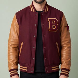 May include: A varsity jacket featuring a burgundy wool body with tan leather sleeves, a letter B patch on the chest, a star patch on the sleeve, and striped ribbed trim at the collar, cuffs, and hem, worn over a black t-shirt and black jeans.