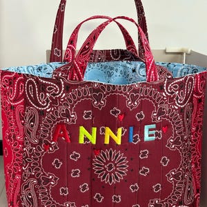 Op de afbeelding: Een gewatteerde draagtas met een donkerrood paisley bandana-patroon en twee handvatten. De naam ANNIE is in kleurrijke letters op de voorkant geborduurd, geaccentueerd door kleine rode hartjes. De binnenvoering is van lichtblauwe paisley-stof.