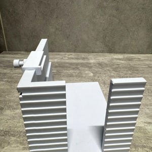 Peut inclure: Un gabarit de guidage en forme de U gris clair, doté d'une base plate et de deux parois latérales parallèles à nervures verticales. Un petit mécanisme à vis métallique réglable est fixé sur le bord supérieur d'une paroi latérale.