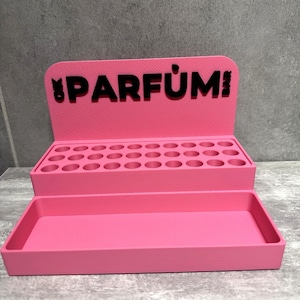 May include: A bright pink tiered display stand featuring a back panel with the black text PARFUM BAR and a small rabbit icon. The upper tier contains three rows of circular cutouts for small bottles, while the lower tier is a rectangular tray.