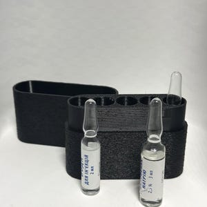 Op de afbeelding: Een zwarte 3D-geprinte opbergdoos met meerdere cilindrische sleuven, met twee doorzichtige glazen ampullen met witte etiketten met tekst in het Cyrillisch. Eén ampul is gelabeld 2 ml en de andere 2,5% 3 ml, met één lege sleuf zichtbaar.