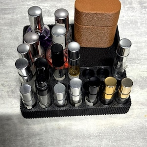 Könnte beinhalten: Ein schwarzes, rechteckiges Organizer-Tablett mit mehreren runden Schlitzen, die verschiedene kleine Glasfläschchen mit silbernen, schwarzen und goldenen Verschlüssen enthalten, sowie ein hohes, strukturiertes, braunes Etui in der hinteren Ecke.
