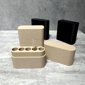 Puede incluir: Una colección de recipientes de almacenamiento ovalados con superficies beige texturizadas y dos recipientes negros sólidos. Un recipiente beige está abierto, revelando cinco ranuras circulares, con un vial de vidrio transparente en una abertura.