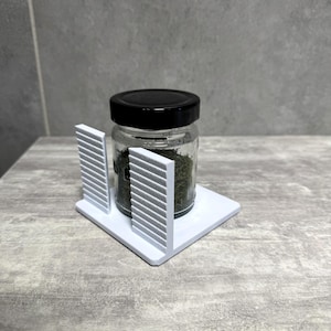 Op de afbeelding: Een witte kunststof organizerstandaard met een platte vierkante basis en twee verticale zijpanelen met horizontale ribbeltexturen. Een kleine doorzichtige glazen pot met een zwarte schroefdop en gedroogde groene kruiden staat ertussen.