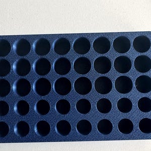 Puede incluir: Una bandeja de almacenamiento rectangular negra con una cuadrícula de cuarenta hendiduras circulares dispuestas en cinco filas y ocho columnas. La superficie de la bandeja tiene un patrón texturizado de líneas cruzadas en toda la cara superior.