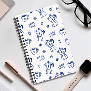 Könnte beinhalten: Ein spiralgebundenes Notizbuch mit weißem Einband und einem wiederkehrenden blauen Muster aus Espressokochern, Kaffeetassen mit Herzen, Kaffeebohnen, kleinen Herzen sowie dem Schriftzug "COFFEE LOVER" in einer einfachen serifenlosen Schrift.