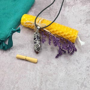 May include: A dark crystal pendant with a silver-toned metal cage featuring floral details, suspended on a black cord. The set includes a yellow honeycomb-textured candle, a small rolled paper scroll with a gold ring, and purple dried flowers.