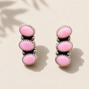 May include: A pair of silver earrings, each featuring three vertically aligned oval pink stones set in serrated metal bezels. The stones are separated by small silver spheres, creating a linear design with a polished finish and a bright pink hue.