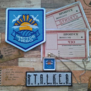 May include: A blue shield-shaped patch with a yellow sun and white birds, a rectangular patch with the text S.T.A.L.K.E.R., a silver pendant necklace, and two rectangular paper passes with Cyrillic text and the word Pripyat printed on them.