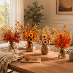 May include: Four glass and ceramic vases filled with autumn floral arrangements featuring orange and yellow sunflowers, small pumpkins, dried foliage, and berries. A single small orange pumpkin sits on a wooden board in the foreground.