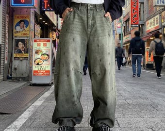 Puede incluir: Pantalones vaqueros de color verde oliva con un corte holgado, un lavado desgastado y sutiles manchas oscuras. Los vaqueros presentan un talle alto, bolsillos laterales y una silueta de pierna ancha. Zapatos de plataforma negros con pinchos.