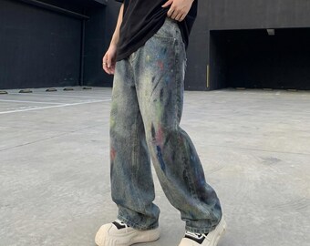 Puede incluir: Pantalones vaqueros de mezclilla azul de corte holgado con un lavado desgastado y un patrón de salpicaduras de pintura multicolor. Los vaqueros tienen un corte recto, diseño clásico de cinco bolsillos y cierre de botones, con zapatillas blancas.