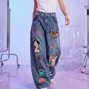 Op de afbeelding: Blauwe spijkerbroek van denim met wijde pijpen en een hoge taille. De broek is versierd met diverse kleurrijke geborduurde patches, waaronder stripfiguren van honden, een kopje, sterren, hartjes en tekst zoals "HA HA", "LUCKY" en "MAFK VISTAGE".