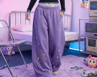 Puede incluir: Pantalones de chándal morados de corte holgado con una cintura elástica que tiene el texto Cailelv Kelin impreso repetidamente. La tela está decorada con varios gráficos de estrellas blancas y amarillas y un gran diseño de estrella en espiral en la pierna derecha.