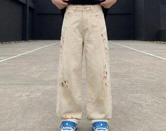 Puede incluir: Pantalones vaqueros de color beige con corte ancho, cintura alta con cierre de botón y salpicaduras de pintura multicolor en los paneles laterales. Los pantalones incluyen bolsillos delanteros y se combinan con zapatillas blancas con cordones azules.