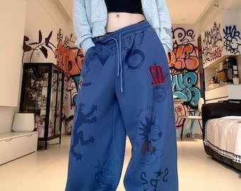 May include: A pair of loose-fitting blue sweatpants featuring various dark blue and red graphic prints, including a cartoon character, the text "SO", and abstract shapes. The sweatpants have a drawstring waist and are paired with a black crop top.