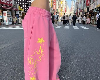 Puede incluir: Pantalones de chándal de color rosa brillante con cintura elástica alta y silueta de pierna ancha. El lateral de la pierna está decorado con un estampado gráfico vertical de varias estrellas amarillas de distintos tamaños en cascada.