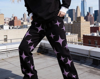Puede incluir: Pantalones de chándal negros con un patrón repetitivo de estrellas moradas de cuatro puntas. Los pantalones tienen un corte relajado, una cintura con cordón ajustable y están confeccionados en tejido de punto. El diseño cubre toda la prenda.