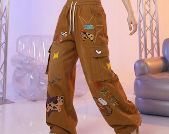 Puede incluir: Pantalones cargo de color marrón con cintura elástica alta y cordones blancos. Los pantalones están decorados con gráficos de perros bordados, estrellas amarillas y el texto "mode sister" y "sister" en una tipografía colorida.