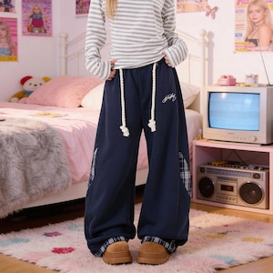 May include: Navy blue wide-leg sweatpants featuring thick white rope drawstrings at the waist, a small white embroidered script logo on the upper left thigh, and blue and white plaid fabric panels visible at the side seams and the pant cuffs.