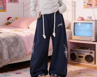 Puede incluir: Pantalones de chándal azul marino de pierna ancha con cordones gruesos de color blanco en la cintura, un pequeño logotipo bordado en blanco en el muslo izquierdo y paneles de tela a cuadros azules y blancos en las costuras laterales y los bajos.