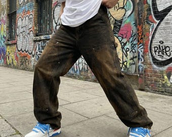Puede incluir: Pantalones vaqueros de color marrón oscuro con efecto lavado al ácido, de corte holgado y relajado, con paneles reforzados en las rodillas, remaches metálicos y bolsillos utilitarios. Se combinan con una camiseta blanca y zapatillas blancas con cordones azules.