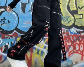 Puede incluir: Pantalones cargo negros con múltiples bolsillos con cremallera, herrajes de metal plateado y correas de tela desmontables adornadas con un patrón repetitivo de pequeñas estrellas rojas. Los pantalones tienen una silueta de pierna ancha y zapatillas blancas.