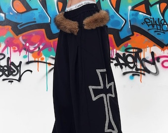 Puede incluir: Pantalones negros de pierna ancha con un borde de piel sintética marrón a lo largo de la cintura y un gran gráfico de cruz en el lateral de la pierna adornado con pedrería plateada. El pantalón tiene un corte de cintura alta y silueta fluida.