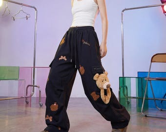 Puede incluir: Una camiseta sin mangas blanca y pantalones cargo negros con cintura de cordón, bolsillos laterales y múltiples apliques de osos de peluche marrones. Una cabeza de oso de peluche grande está unida al bolsillo de la pierna izquierda, con parches y lazos.