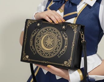 Puede incluir: Un bolso de hombro rectangular de color negro con diseños dorados impresos de una luna creciente, estrellas y patrones geométricos. El bolso incluye una correa larga para el hombro y detalles metálicos dorados, con el texto Magic Book en el frente.