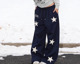 May include: Navy blue wide-leg sweatpants featuring white fuzzy star appliques and a drawstring waist. The text "Happy homing" is embroidered in white near the right hip. The pants are paired with a light gray zip-up hoodie and white sneakers.