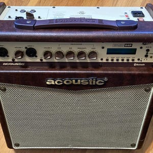 Puede incluir: Un amplificador de la marca Acoustic con un exterior texturizado de color marrón oscuro y una rejilla de altavoz color crema. El panel de control superior cuenta con múltiples perillas para volumen y ecualización, pantalla y asa.