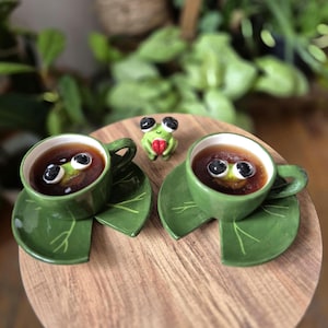 Könnte beinhalten: Zwei grüne Keramiktassen mit passenden Untertassen in Seerosenblattform. Jede Tasse enthält eine dunkle Flüssigkeit und eine kleine Froschfigur mit großen schwarzen Augen. Eine winzige Froschfigur mit einem roten Herz steht zwischen den Tassen.