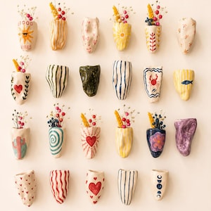 Peut inclure: Une collection de vingt-trois petits vases muraux en céramique aux formes irrégulières disposés en grille. Chaque récipient présente des motifs peints à la main, notamment des soleils, des cœurs, des rayures, des fleurs et un crabe.