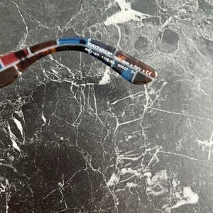 Puede incluir: La patilla interior de unas gafas de sol con estampado de carey y detalles en azul y rojo. El texto impreso dice WAYFARER, RB 2140 1108/85 50 22 3N y Hand Made in Italy. La patilla es curva y presenta un acabado brillante con letras blancas.