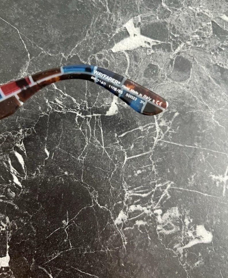 Puede incluir: La patilla interior de unas gafas de sol con estampado de carey y detalles en azul y rojo. El texto impreso dice WAYFARER, RB 2140 1108/85 50 22 3N y Hand Made in Italy. La patilla es curva y presenta un acabado brillante con letras blancas.
