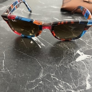 Puede incluir: Gafas de sol Ray-Ban con montura multicolor que presenta un patr&oacute;n de cuadrados azules, naranjas y marr&oacute;n oscuro. Las lentes son de color marr&oacute;n oscuro y la montura incluye detalles de metal plateado en las esquinas. Se ve un estuche de cuero beige.